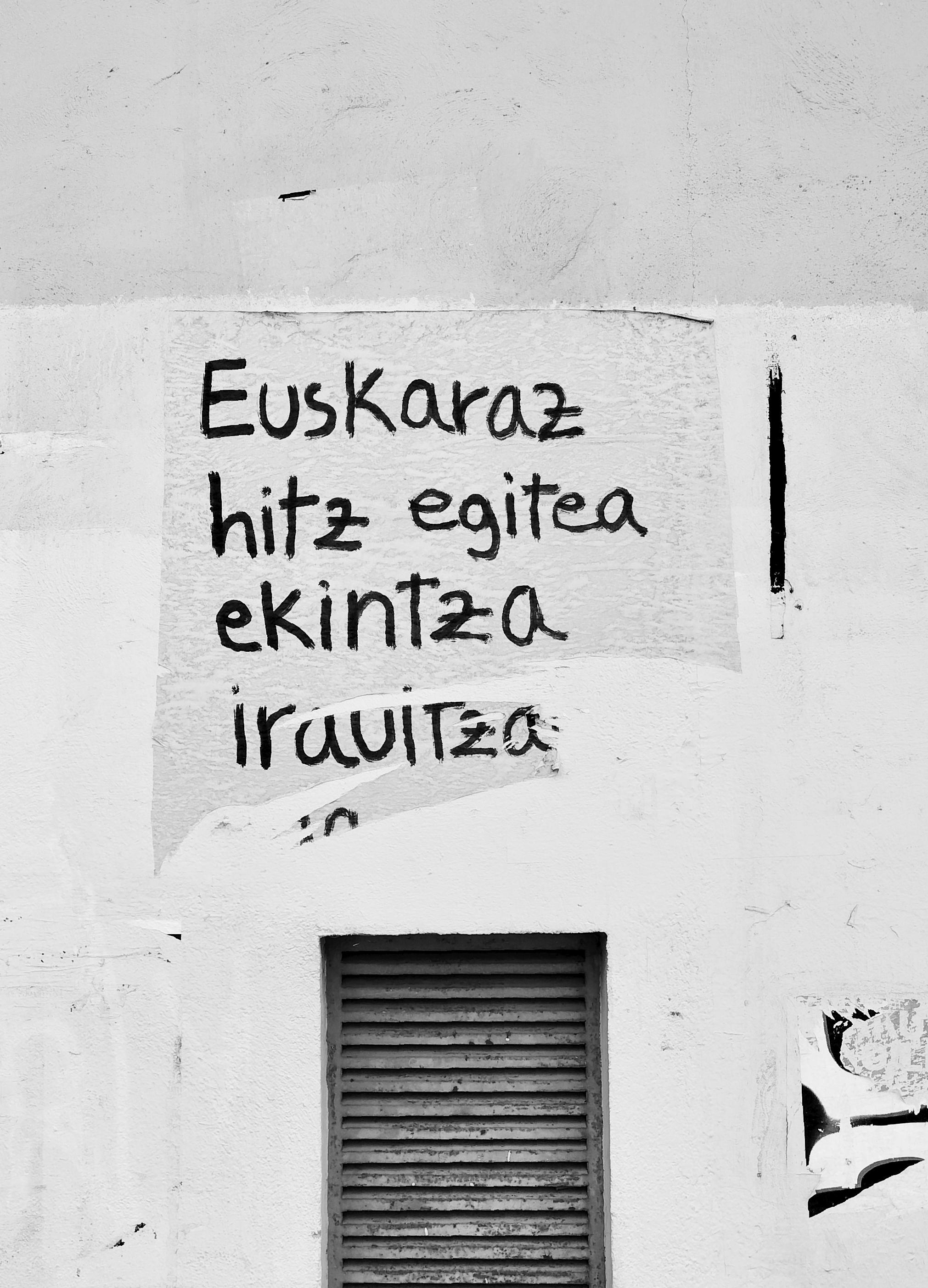 Horma batean kolatutako kartel urratu xamar bat agertzen da irudian. Bertan, eskuz idatzita, haixe irakurri diateke: «Euskaraz hitz egitea ekintza iraultza...».