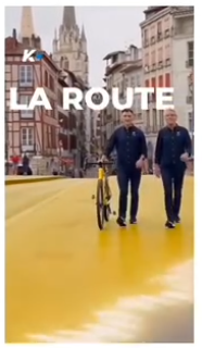 ETB Kirolakek Tourra iragartzeko Baionan grabatu duen iragarkiaren pantaila irudia. Bi aurkezle ageri dira, oinez doazela Baionako zubi batean zehar, katedrala ikusten dela, eta LA ROUTE idatzita dagoela.