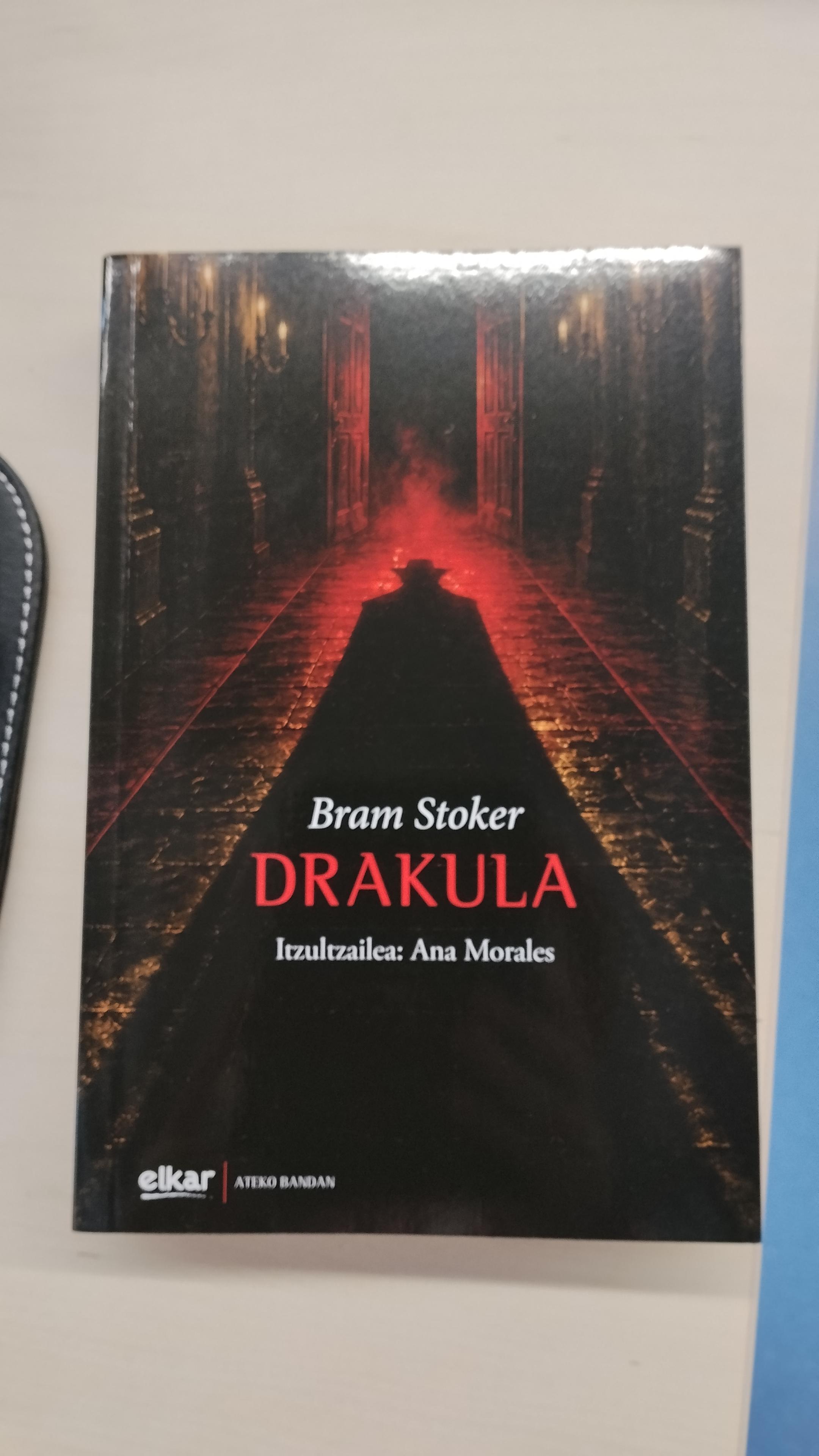 Drakula nobelaren euskarazko bertsioaren azala. Karraju ilun batean, figura beltz baten itzal iluna hedatzen da, distira gorri baten kontra. Horma beltzetan kandela batzuk daude piztuta. Figuraren gainean, letra zurietan "Bram Stoker" jartzen du, azpian letra gorriz, "Drakula", eta horren azpian, letra zuriz, "Itzultzailea: Ana Morales"